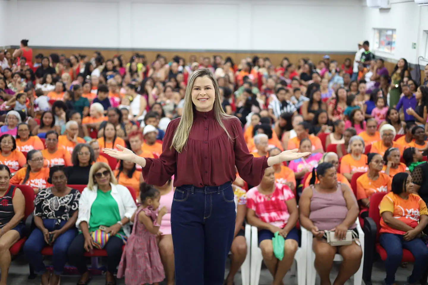 Mulher de blusa vinho em pé diante de um auditório lotado