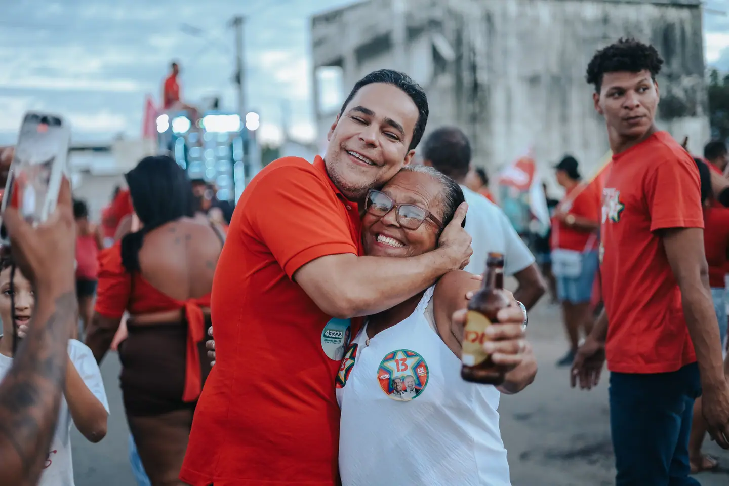 Homem de camisa vermelha abraça uma senhora sorridente na rua